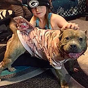 Rockie is registered to the contest to win money with this photo: dog, pet, camouflage_shirt, person, cap, carpet, indoor, home, furniture, smiling, animal, canine, friendly, happy, hand, casual, clothing, floor, relaxed, domestic