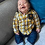 baby, child, smiling, plaid_shirt, blue_jeans, sneakers, couch, pillow, indoor, happy, seated, infant, casual_clothing, cozy, portrait, cute, young, small_hands, comfortable, relaxed