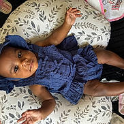Miyori is registered to the contest to win money with this photo: baby, infant, child, portrait, smiling, lying_down, blue_outfit, bonnet, dress, cushion, blanket, baby_bottle, bib, nursery, indoor, cozy, patterned_fabric, dark_skin, bare_feet, happy