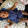 baby, infant, child, portrait, smiling, lying_down, blue_outfit, bonnet, dress, cushion, blanket, baby_bottle, bib, nursery, indoor, cozy, patterned_fabric, dark_skin, bare_feet, happy