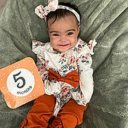 Isabella joined the competition — help win amazing prizes! baby, infant, child, smiling, headband, bow, floral_clothing, orange_pants, milestone_card, blanket, soft_texture, cute, happy, portrait, lying_down, young_child, indoors, person, footwear, fashion