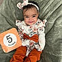 baby, infant, child, smiling, headband, bow, floral_clothing, orange_pants, milestone_card, blanket, soft_texture, cute, happy, portrait, lying_down, young_child, indoors, person, footwear, fashion