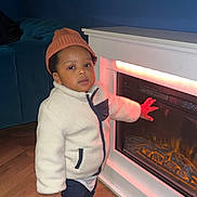 Kenan participe au concours pour gagner de l'argent avec cette photo : beanie, blue_wall, child, couch, cozy, curious, electric_fire, fireplace, flooring, furnishing, hand_on_glass, home, indoor, jacket, lighting, portrait, sneakers, standing, toddler, wood_floor