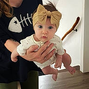Eden participe au concours pour gagner de l'argent avec cette photo : baby, child, headband, bow, adult_hand, person, indoor, floor, wooden_floor, white_wall, handrail, long_sleeve, curious, wide_eyes, holding, clothing, black_shirt, fishbone_design, human, portrait