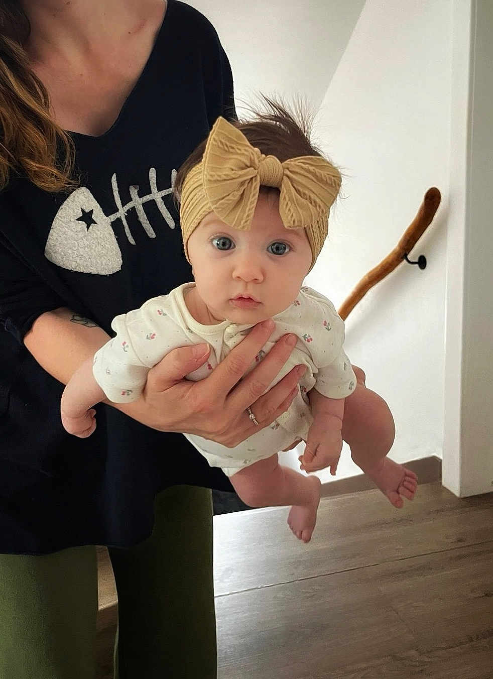 Eden participe au concours pour gagner de l'argent avec cette photo : baby, child, headband, bow, adult_hand, person, indoor, floor, wooden_floor, white_wall, handrail, long_sleeve, curious, wide_eyes, holding, clothing, black_shirt, fishbone_design, human, portrait