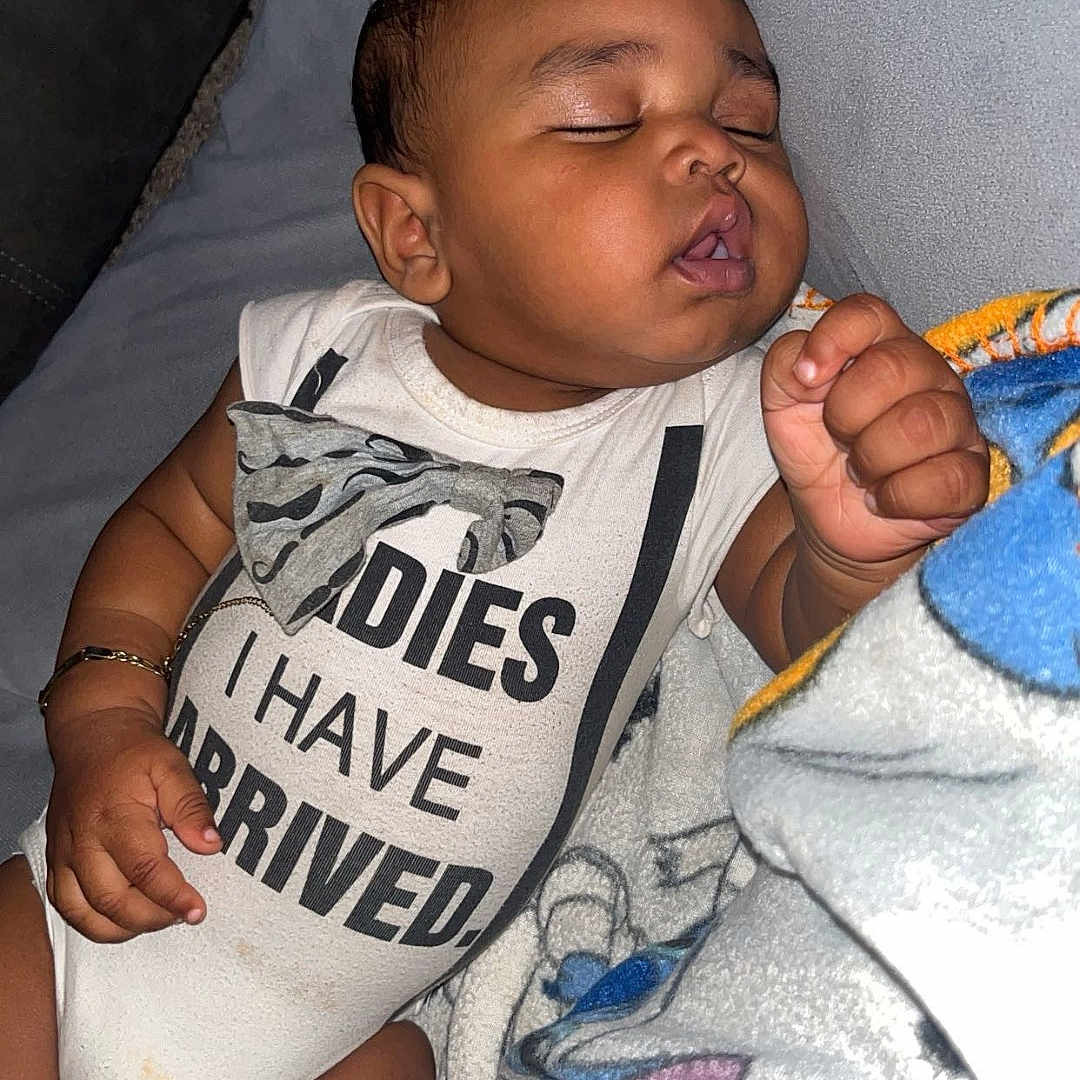 Kycen is registered to the contest to win money with this photo: baby, sleeping, onesie, text_on_clothing, blanket, indoor, child, infant, cute, peaceful, resting, soft_texture, closeup, portrait, person, clothing, hand, face, nap, cozy