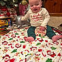 baby, child, christmas, holiday, blanket, toy, present, decorations, smiling, festive_clothing, indoor, floor, wood_paneling, carpet, face, person, sitting, cute, seasonal, happy
