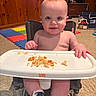 baby, infant, high_chair, tray, food, smile, face, eyes, carpet, wood_paneling, play_mat, toy, socks, shoes, rosy_cheeks, indoor, messy_eating, cute, portrait, child