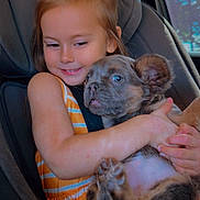 Chanel is registered to the contest to win money with this photo: girl, child, puppy, dog, pet, hugging, smiling, car, car_seat, portrait, indoors, sitting, bow, dress, brown_fur, blue_eyes, animal, family, cute, photography