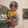 Aiden is registered to the contest to win money with this photo: toddler, child, baby, sunglasses, yellow_bib, holding, adult_hand, pajamas, gray_pajamas, dinosaur_print, indoor, kitchen, cabinet, countertop, person, portrait, fashion, eyewear, cute, funny