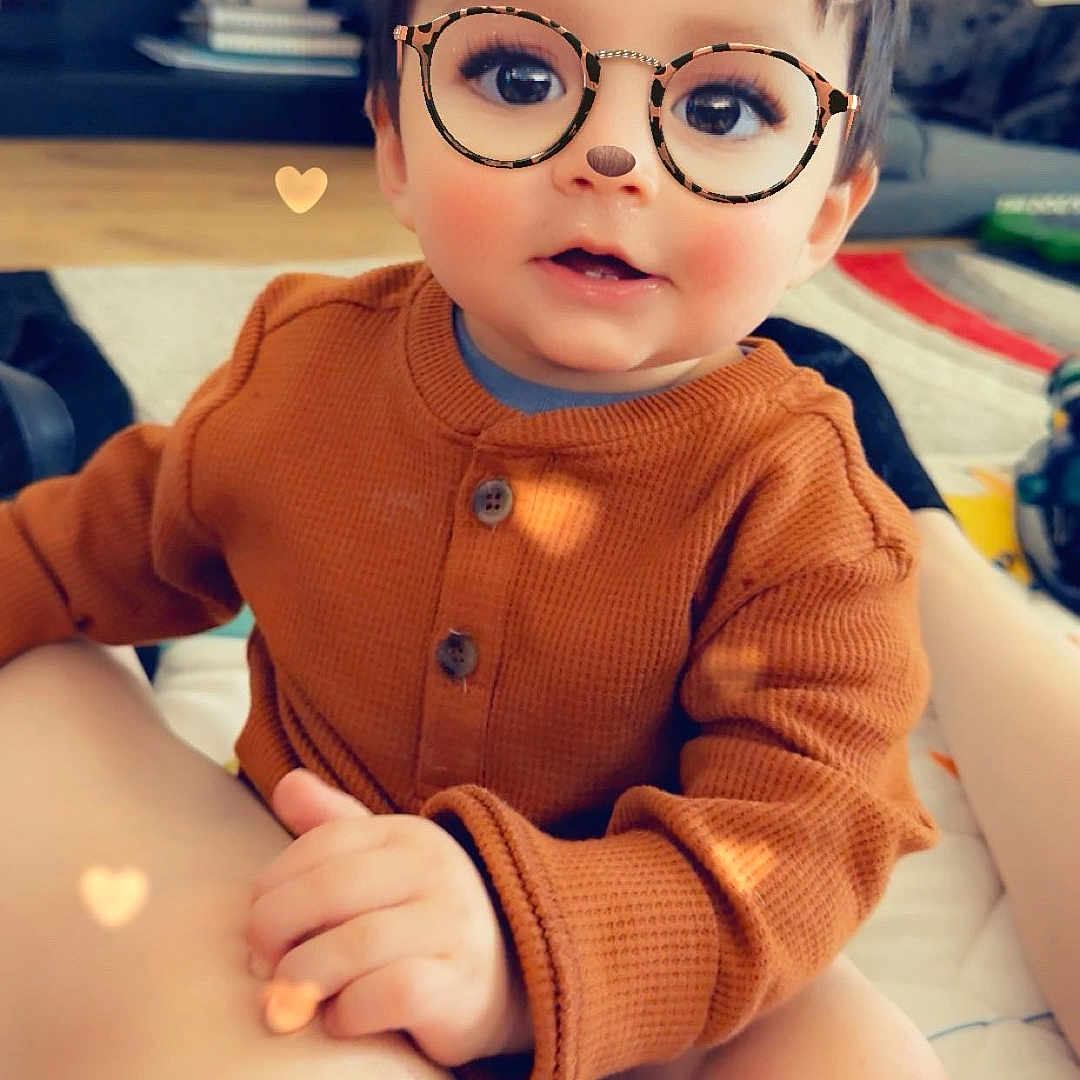 Aiden is registered to the contest to win money with this photo: bear_ears_filter, brown_sweater, carpet, child, cozy, cute, digital_filter, face, floor, furniture, glasses_filter, hand, heart_bokeh, indoors, legs, playful, portrait, sitting, smile, toddler