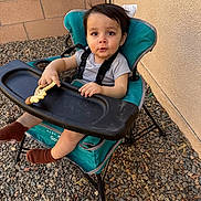 Aiden is registered to the contest to win money with this photo: toddler, child, high_chair, teether, outdoor, rocks, wall, baby, seat, toy, person, footwear, brown_socks, clothing, expression, face, hair, curious, looking_at_camera, seatbelt