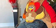 Aiden joined the competition — help win amazing prizes! baby, child, turkey_outfit, jeans, socks, indoor, person, adult_hand, plant, flower_painting, vase, feathers, curious, seated, colorful_clothing, portrait, home_interior, cute, casual_wear, holding