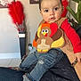 baby, child, turkey_outfit, jeans, socks, indoor, person, adult_hand, plant, flower_painting, vase, feathers, curious, seated, colorful_clothing, portrait, home_interior, cute, casual_wear, holding