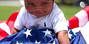 Santana joined the competition — help win amazing prizes! child, toddler, curly_hair, smile, american_flag, grass, outdoor, white_shirt, playful, baby, face, person, summer, daylight, crawling, cheeky, cute, happy, fun, patriotic