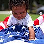 child, toddler, curly_hair, smile, american_flag, grass, outdoor, white_shirt, playful, baby, face, person, summer, daylight, crawling, cheeky, cute, happy, fun, patriotic