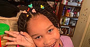 Kimani is registered to the contest to win money with this photo: braids, casual, child, cozy, furniture, girl, hair_clips, hand_gesture, happy, home, indoor, living_room, person, playful, portrait, purple_shirt, shelf, smile, toys, unicorn