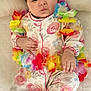 Shams is registered to the contest to win money with this photo: baby, infant, flower_crown, lei, floral_onesie, pajamas, cute, lying_down, blanket, puffed_cheeks, hand, footie_pajamas, colorful, soft_lighting, indoors, portrait, adorable, sleepwear, flower_accessory, baby_girl
