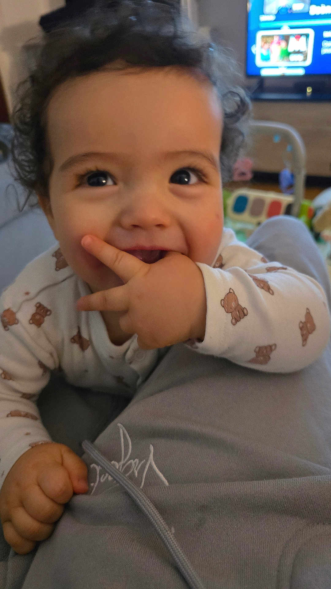 Wyatt participe au concours pour gagner de l'argent avec cette photo : baby, child, curly_hair, onesie, teddy_bear_pattern, hand, fingers, smile, happy, indoor, living_room, television, couch, person, closeup, cute, playful, infant, face, eyes