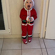 Juliette a rejoint le concours — aidez-le/la à gagner de superbes lots ! child, santa_claus, costume, indoor, tile_floor, door, wall, tongue_out, playful, holiday, festive, red_clothing, hat, footwear, person, cute, holding_object, smiling, young_child, standing
