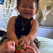 Athena is registered to the contest to win money with this photo: toddler, child, smiling, barefoot, toy, bed, indoor, plant, vines, necklace, black_top, curly_hair, window, home, happy, portrait, cute, person, room, decor