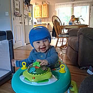 Onyx is registered to the contest to win money with this photo: baby, blue_hat, chair, child, couch, daylight, dining_table, footwear, furniture, happy, home, indoor, kitchen, living_room, person, playtime, smiling, toy, walker, wooden_floor