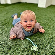 Mael a rejoint le concours — aidez-le/la à gagner de superbes lots ! baby, infant, grass, pacifier, cardigan, blue_eyes, smile, happy, child, outdoor, toy, lying_down, cute, young_child, portrait, blond_hair, clothing, person, nature, daylight