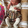 baby, blanket, child, colorful_background, crocheted_blanket, cute, furniture, hand_in_mouth, home, indoors, infant, newborn, patterned_blanket, plaid_skirt, portrait, resting, sitting, small_person, soft_texture, white_outfit