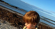 Dawson is registered to the contest to win money with this photo: beach, blue_sky, boy, calm, casual_clothing, child, coast, daylight, landscape, nature, outdoor, portrait, profile, rocks, sand, sea, shoreline, sky, sunlight, young