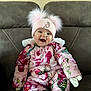 baby, child, smiling, pink_hat, floral_snowsuit, mittens, couch, indoor, happy, cute, winter_clothing, baby_face, seated, cozy, warm_clothes, infant, portrait, funny_hat, pompoms, home