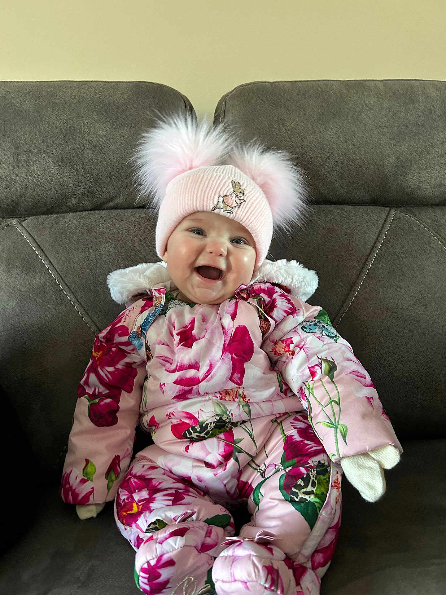 Lylah is registered to the contest to win money with this photo: baby, child, smiling, pink_hat, floral_snowsuit, mittens, couch, indoor, happy, cute, winter_clothing, baby_face, seated, cozy, warm_clothes, infant, portrait, funny_hat, pompoms, home