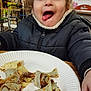 blurred_background, casual, child, chocolate, clothing, crepes, expression, face, food, funny_face, happy, indoor, jacket, meal, person, plate, sitting, table, tongue, young