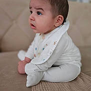 Alper joined the competition — help win amazing prizes! baby, infant, child, white_clothing, bib, sitting, couch, beige, soft_fabric, curious, side_view, indoors, young_child, cross_legged, face, expression, cute, small_hands, head, portrait