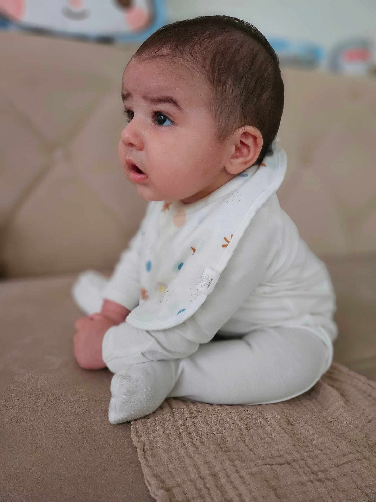 Alper joined the competition — help win amazing prizes! baby, infant, child, white_clothing, bib, sitting, couch, beige, soft_fabric, curious, side_view, indoors, young_child, cross_legged, face, expression, cute, small_hands, head, portrait