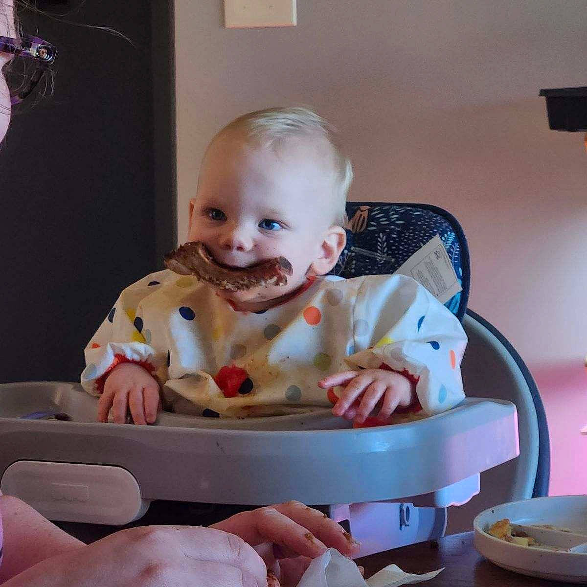 William is registered to the contest to win money with this photo: baby, baby_products, baby_toddler_clothing, chair, cheek, child, drinkware, eating, fun, happy, mouth, person, pink, plate, room, serveware, sitting, smile, sweetness, tableware