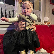 Léonie a rejoint le concours — aidez-le/la à gagner de superbes lots ! baby, child, person, adult_hand, headband, bow, black_skirt, golden_boots, red_couch, window, festive_decor, indoor, cozy, warm_lighting, toy_dog, curious_expression, holding, sitting, clothing, portrait