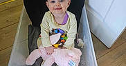 Lillie is registered to the contest to win money with this photo: baby, black_pillow, child, clothing, cushion, drool, face, floor, happy, head, indoors, person, pink, plastic_container, purple, seated, smiling, stuffed_animal, toy, wooden_floor