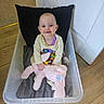 baby, child, smiling, plastic_container, stuffed_animal, toy, cushion, floor, wooden_floor, clothing, indoors, drool, happy, person, face, head, seated, pink, purple, black_pillow