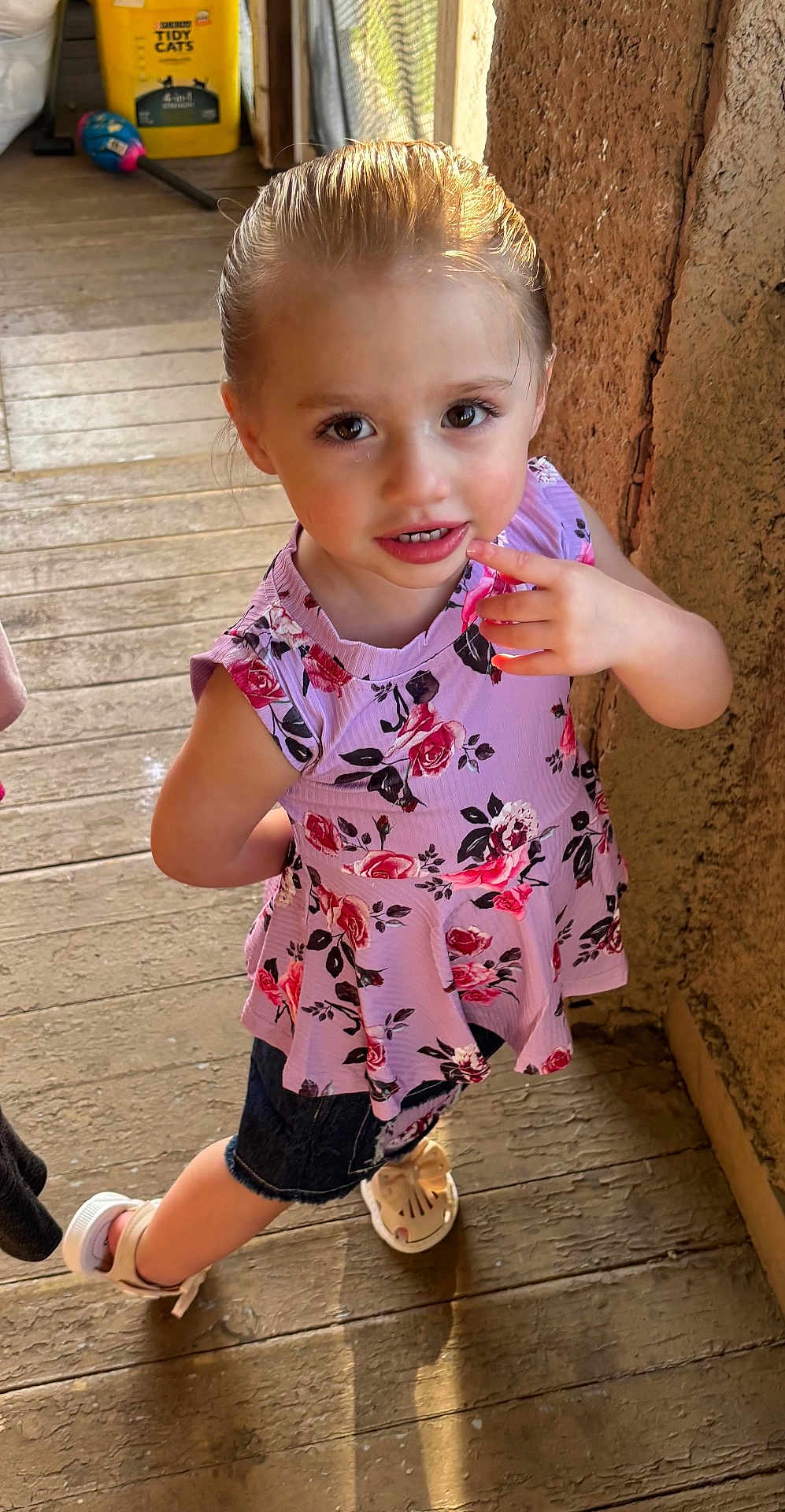 Lilianna is registered to the contest to win money with this photo: child, girl, floral_top, denim_shorts, sandal, wooden_floor, wall, finger_on_mouth, curious_expression, natural_light, indoors, standing, brown_hair, big_eyes, summer_clothing, casual, cute, young, portrait, person