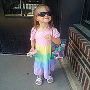 Lilianna joined the competition — help win amazing prizes! child, toddler, girl, sunglasses, rainbow_dress, pink_shirt, colorful_clothing, toy, bag, sandals, standing, outdoor, doorway, brick_wall, sidewalk, portrait, fashion, smiling, sunlight, playful