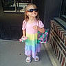 child, toddler, girl, sunglasses, rainbow_dress, pink_shirt, colorful_clothing, toy, bag, sandals, standing, outdoor, doorway, brick_wall, sidewalk, portrait, fashion, smiling, sunlight, playful