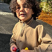 Rakan a rejoint le concours — aidez-le/la à gagner de superbes lots ! child, curly_hair, toy, outdoor, sunlight, face, smile, sweater, hands, play, person, casual_clothing, closeup, daylight, childhood, happy, young, cute, shadow, street