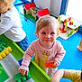 toddler, child, playing, building_blocks, toy, indoor, table, colorful, pink_shirt, blonde_hair, kids, playroom, window, daylight, happy, smiling, person, curly_hair, plastic_blocks, wooden_toy