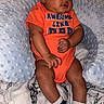 baby, infant, onesie, orange_clothing, socks, pillow, bed, marble_pattern, child, cute, relaxed, indoor, person, skin, head, legs, arms, comfort, text_on_clothing, sleepwear