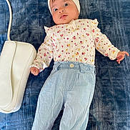 Ayra is registered to the contest to win money with this photo: baby, infant, child, headband, bow, floral_clothing, long_sleeve, pants, blue_pants, white_purse, blanket, blue_blanket, lying_down, cute, wide_eyes, hand, face, person, soft_texture, indoors