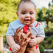 Austin is registered to the contest to win money with this photo: baby, child, smiling, drool, hands, outdoor, greenery, curly_hair, cute, portrait, person, white_shirt, red_clothing, happy, skin, holding, nature, daylight, infant, closeup
