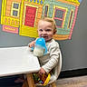 Maverick is registered to the contest to win money with this photo: toddler, child, sippy_cup, toy, high_chair, table, indoor, wall_mural, colorful, smiling, blond_hair, white_clothing, brown_pants, wooden_floor, seat, playful, cute, portrait, happy, person