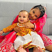 Divit is registered to the contest to win money with this photo: child, baby, smiling, traditional_clothing, red_dress, yellow_outfit, couch, indoor, portrait, happy, face_paint, barefoot, sitting, family, cute, celebration, toddler, cultural, embrace, joy