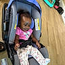 baby, car_seat, infant, pink_shirt, pajamas, baby_bottle, wooden_floor, indoor, child, hair_puffs, clothing, seat_belt, toy, floor, person, baby_gear, casual, cute, relaxed, smiling