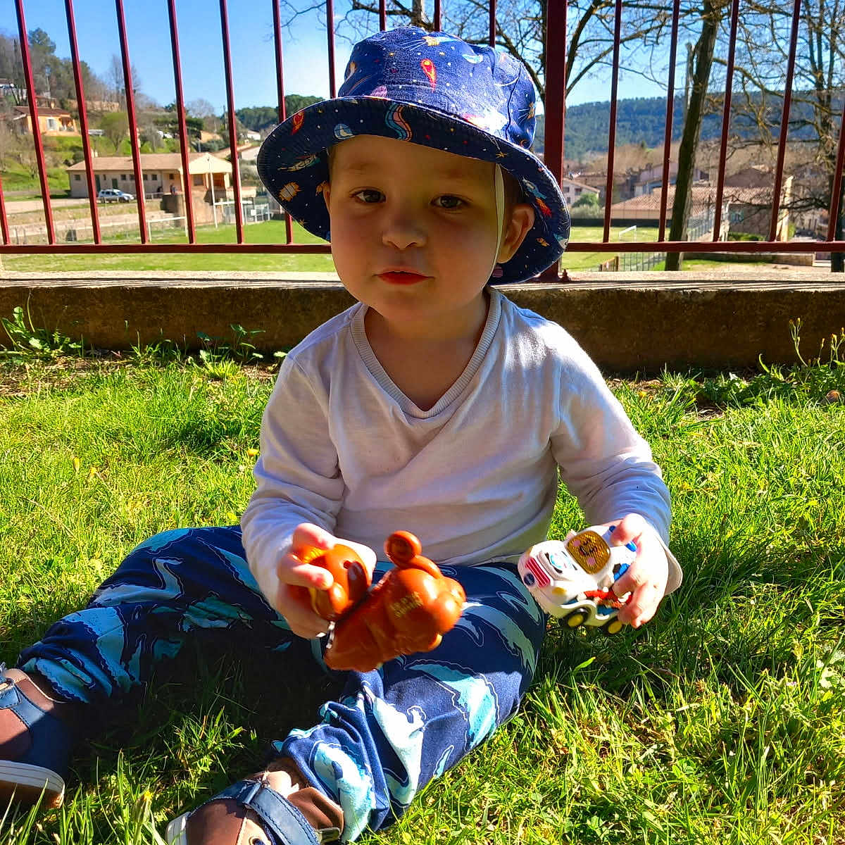 Tarik a rejoint le concours — aidez-le/la à gagner de superbes lots ! blue_sky, bucket_hat, casual_clothing, child, clouds, daylight, fence, grass, hat, nature, outdoor, pants, person, playing, shoes, sunlight, toddler, toy, toy_animal, toy_car